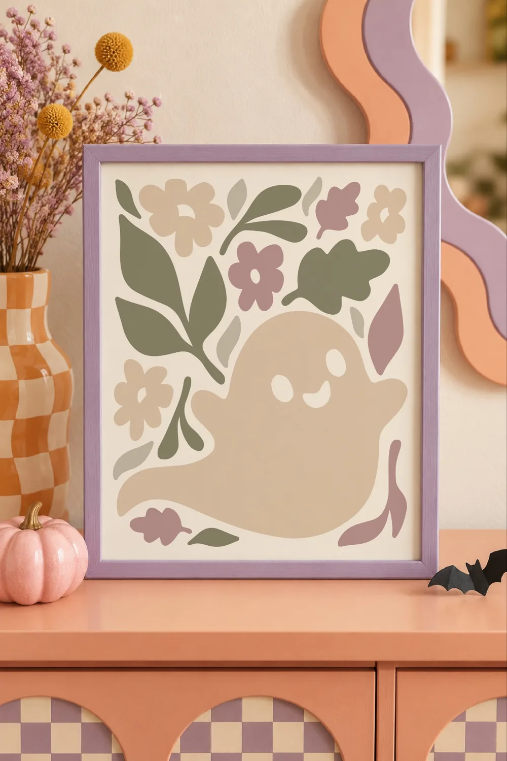 Lavender framed print of a smiling beige ghost with sage leaves and mauve flowers on a peach dresser with Halloween decor