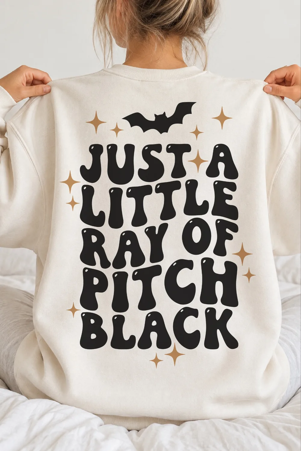 Back of a cream sweatshirt on a woman sitting on a bed, with bubbly black text, a bat and gold sparkle stars