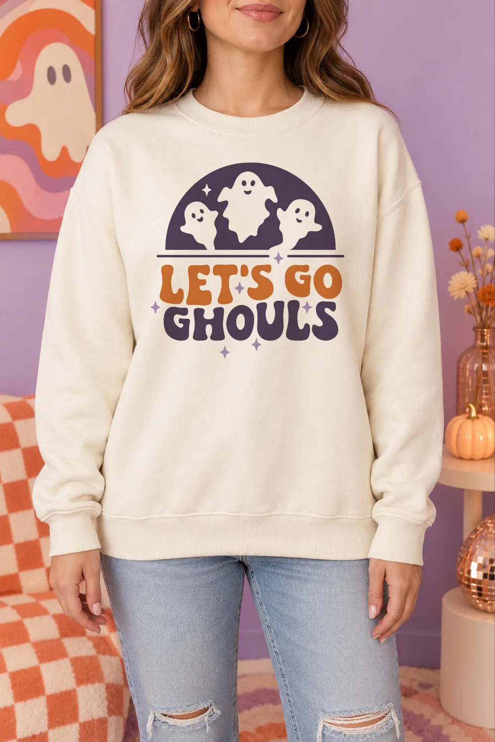 Cream sweatshirt with three smiling ghosts in a purple arch above orange and purple Let's Go Ghouls text, in a lilac room
