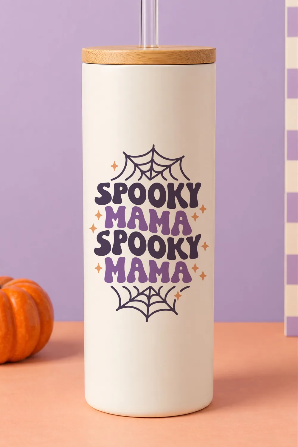 Cream tumbler with bamboo lid and straw, Spooky Mama text with spider webs and stars, beside a pumpkin on a purple backdrop