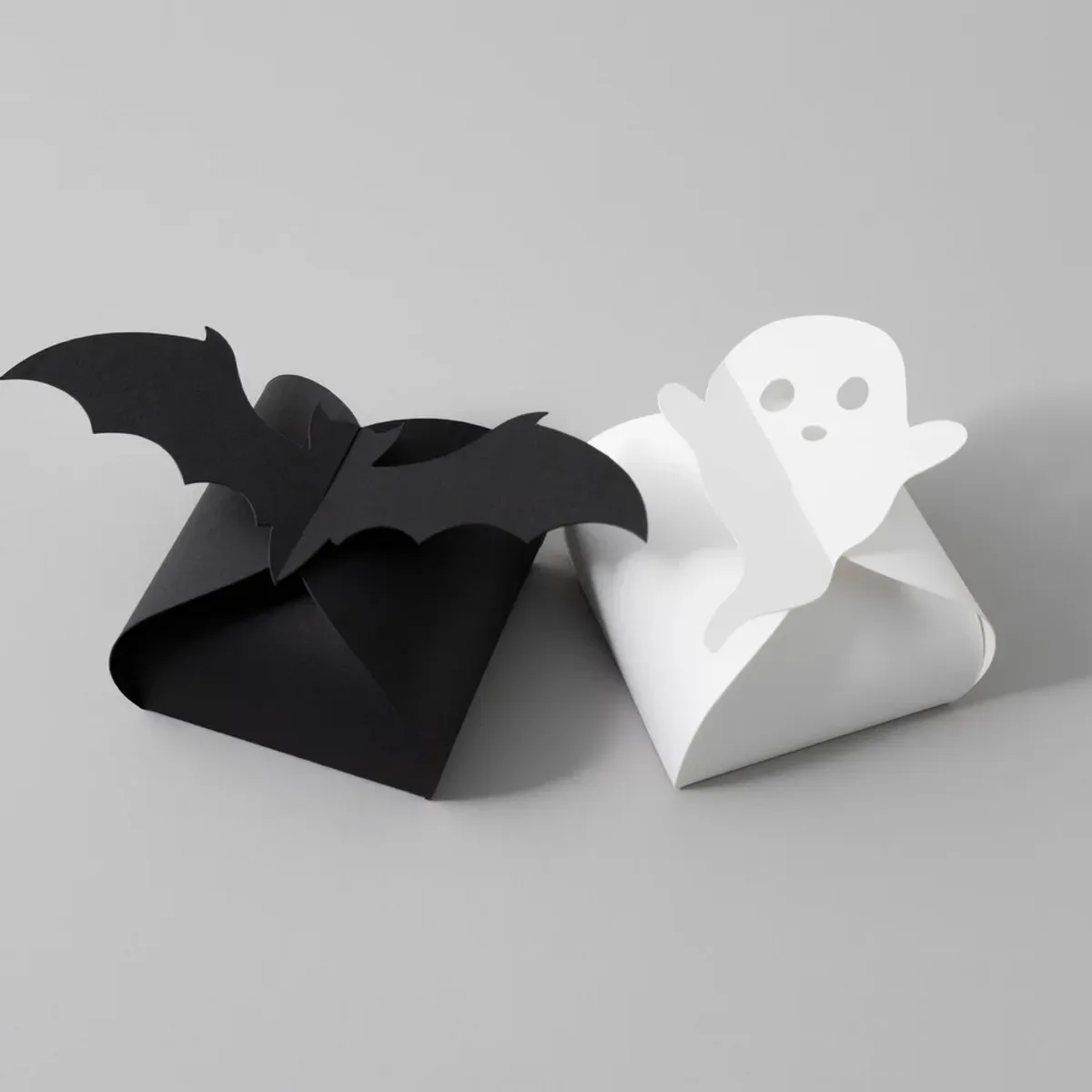 Black cardstock treat box topped with a bat beside a white cardstock treat box topped with a ghost on a gray background